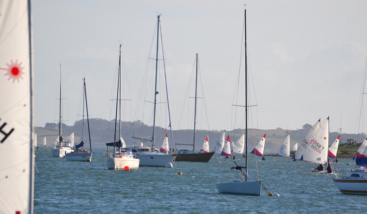 A variety of yachts on moorings with topper dinghies in the background racing on a windy day.