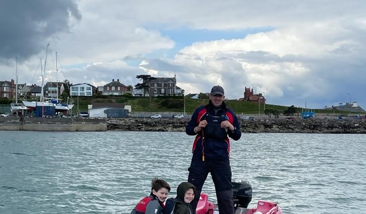 David is standing in a club safety boat on the water with the Bangor shoreline in the background and two volunteers in the front of the shop.