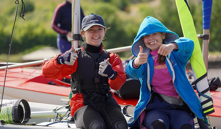 Two junior sailors on a sunny day, smiling and with thumbs up, sat on a boat on shore.