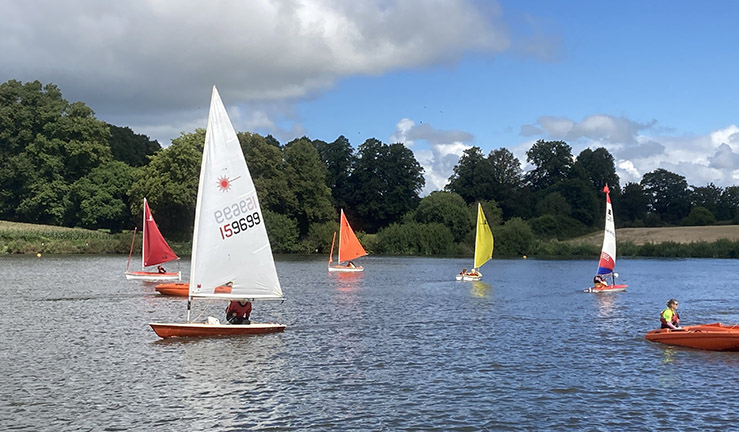 Five dinghies and a RIB on the water in the sunshine at Gresford SC giving visitors from Pride Youth Games an opportunity to experience sailing.