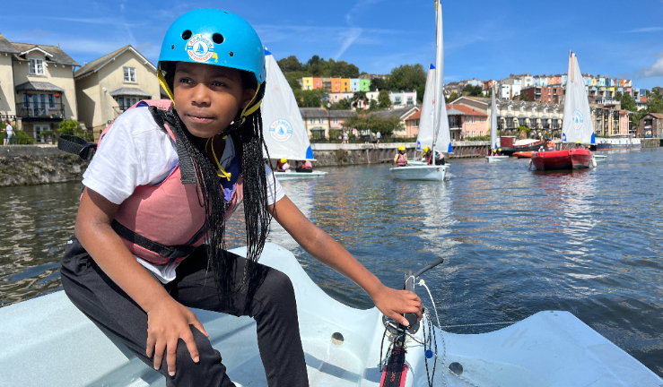 An instructor in a dinghy with four young people wearing helmets and buoyancy aids, all smiling and one giving a thumbs-up