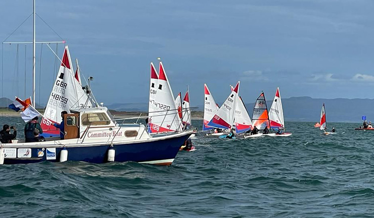 Variety of around a dozen junior dinghies with colourful sails on a startline with the committee boat in the foreground, on a sunny day in Cardigan Bay with mountains in the background, Welsh Youth & Junior Championships 2023.