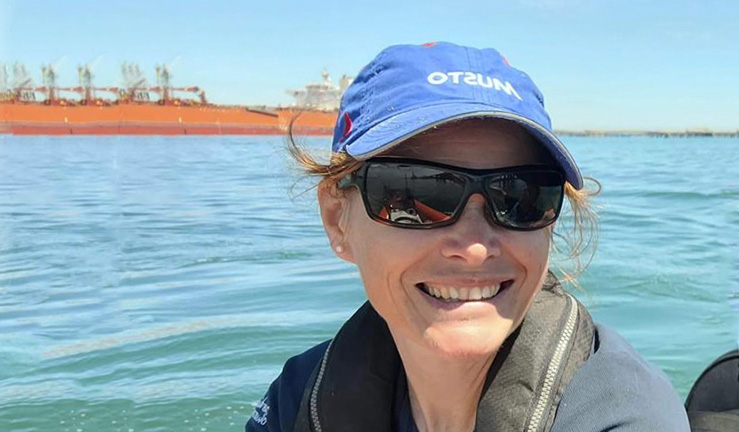 Headshot of RYA CW Club Development Officer Hester Walker smiling for the camera on a sunny day in sunglasses and blue cap with sea and container ship in the background.