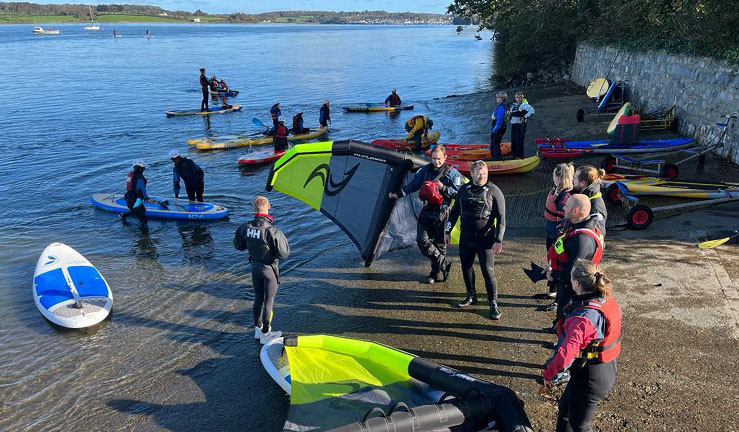 Group of people smiling for the camera outside Plas Menai National Outdoor Centre with RYA feather flags and sunshine for The Big Weekend for clubs, training centres and instructors.