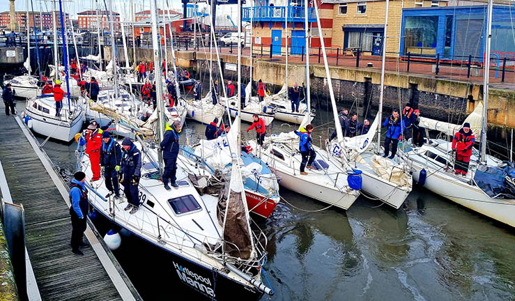 A dozen yachts with sailors on board waiting in sea lock at Hartlepool, buildings on one side, pontoon the other.