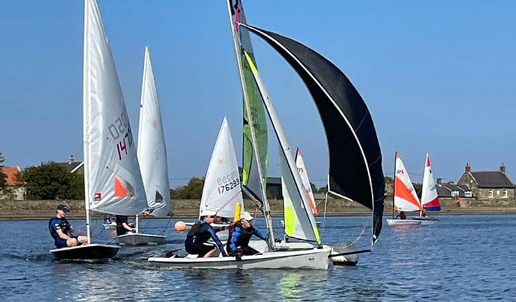 A lake full of colourful youth and junior sailing dinghies racing on a grey day in light winds at Ripon Sailing Club for the North East & Yorkshire Youth Traveller Series 2023.