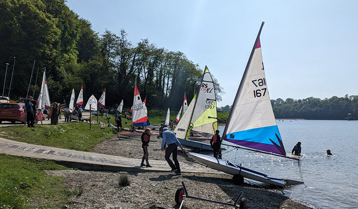 Variety of junior dinghies with colourful sails launching rigging on shore at a lake on a sunny day for a Dragon Series event 2023.