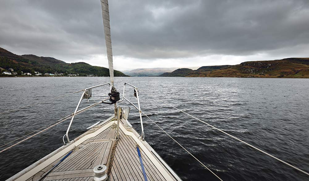 Sloop rigged modern yacht with wooden teak deck sailing on a cloudy day. A view from the deck to the bow. Panoramic view of the rocky shores of Kyles of Bute from the water. Bute island, Scotland, UK