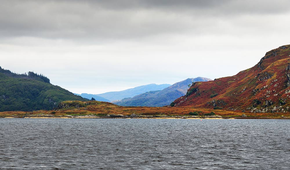 Panoramic view of the rocky shores of Kyles of Bute from the water. Hills and mountains in the background. Dark storm sky. Bute island, Firth of Clyde, Scotland, UK