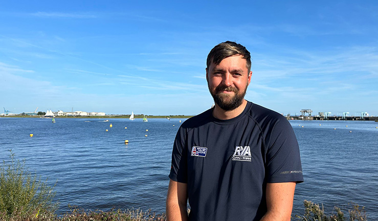 RYA Cymru Wales Events Officer Sam Thomas smiling for the camera in blue T shirt with British Youth Sailing / RYA Cymru Wales logos, in the sunshine with a background of blue sky and blue water with sailing boats.