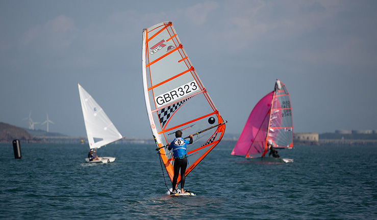 A windsurfer and two dinghies on the water in the sunshine.