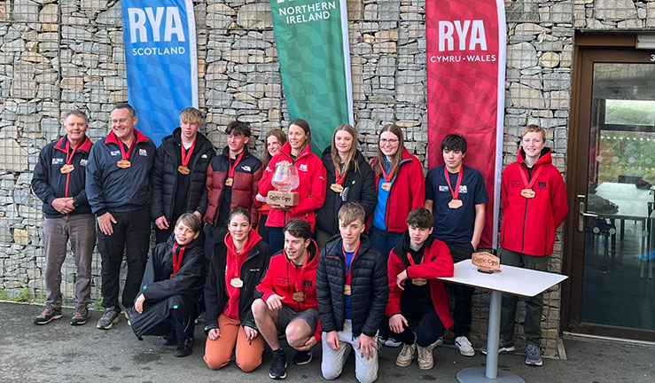 Celtic Cup winning Team Wales sailors with coach Sarah McGovern holding an impressively large glass bowl trophy! Mostly all wearing red, stood in front of the three RYA feather flags for the Home Nations of Scotland, N Ireland and Wales - blue, green and red.