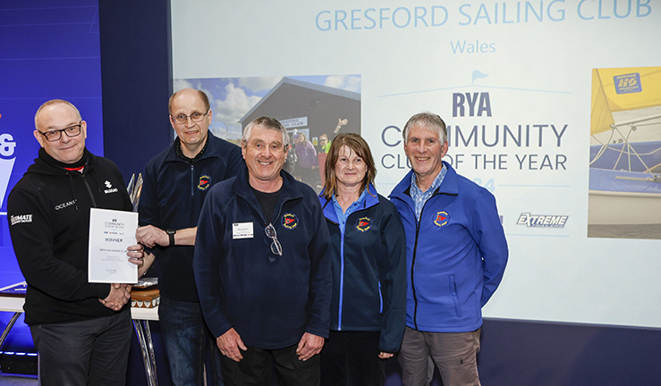Five members of Gresford Sailing Club on stage at the RYA Dinghy & Watersports Show to collect their RYA Community Club of the Year Award certificate, with big screen announcing the award behind them.
