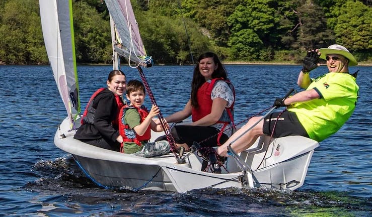 A sailing dinghy with two women and a boy smiling and a helm in a bright yellow T-shirt waving for the camera at a Merthyr Tydfil SC Discover Sailing Day.