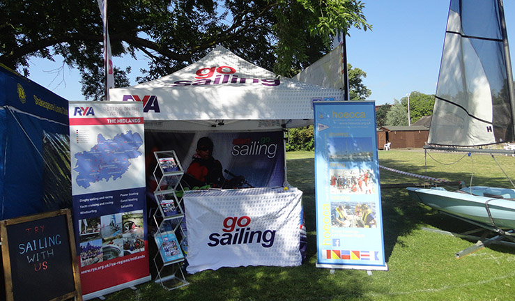 Equipment set out in a green field on a sunny day from one of the RYA Midlands event trailers - including gazebo with 'go sailing' graphic, notice boards and a dinghy on a swivel stand.